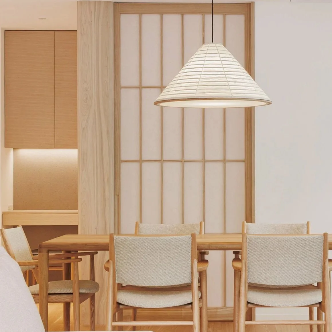 Dining area with wooden table, beige upholstered chairs, a geometric hanging pendant light, and a shoji-style wooden frame on the wall.