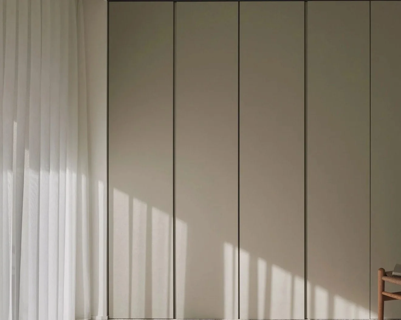 A plain closet with multiple beige-colored doors, partly covered by sheer white curtains with sunlight casting shadows on it.