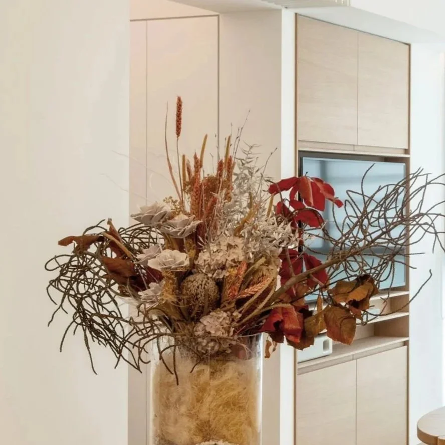Dried flower arrangement in a beige vase inside a modern room with light wood cabinets and a mounted television.