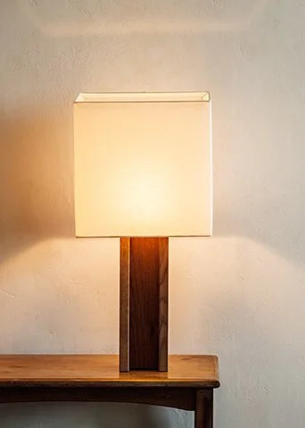 A table lamp with a square off-white lampshade on a wooden table against a plain light-colored wall.