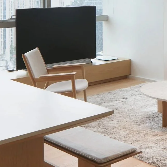 Modern living room with a large flat-screen TV on a wooden stand, a white armchair, a white table with a bench, large window with city view, beige rug, and minimalist decor.