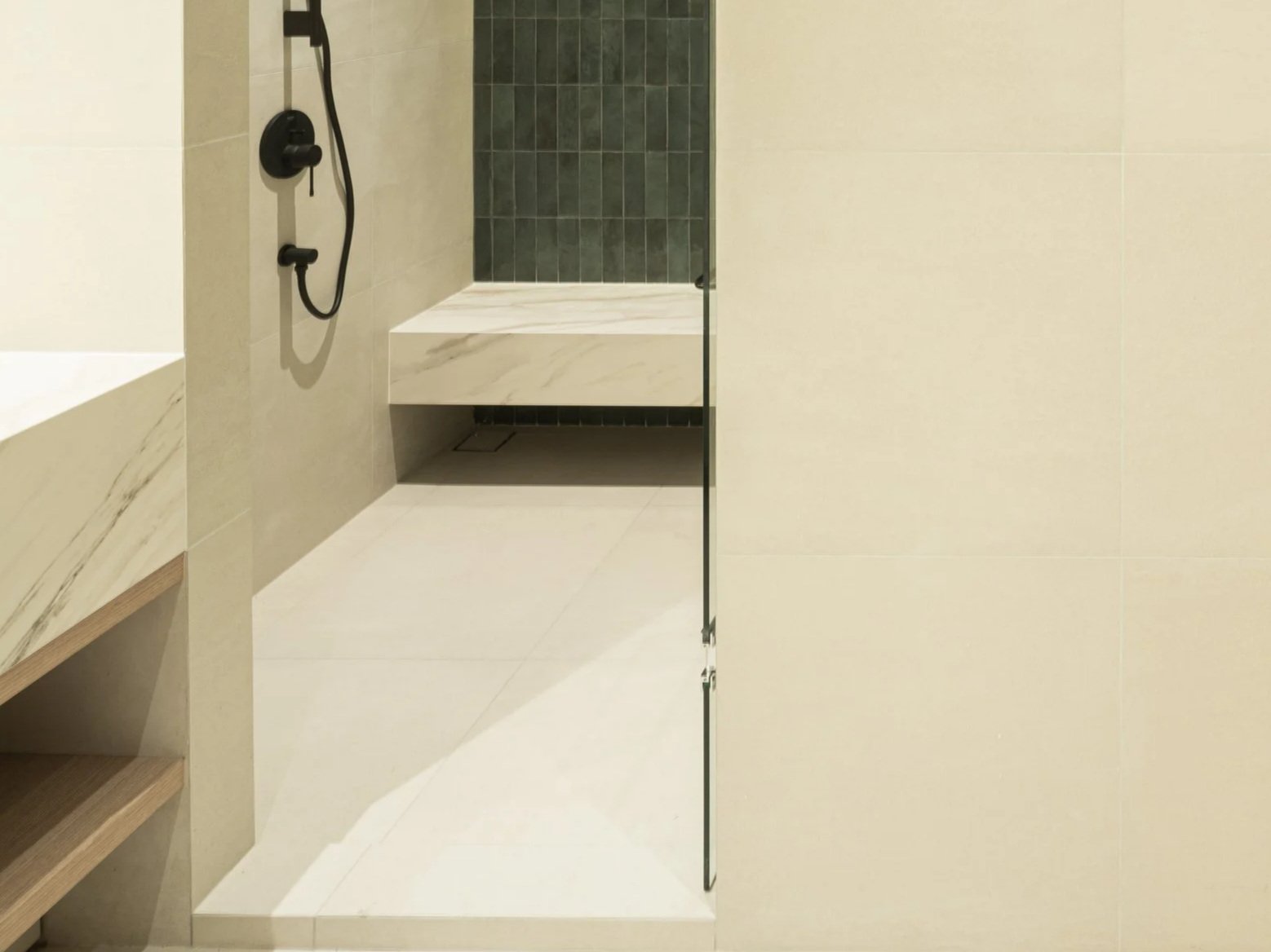 A modern bathroom shower area with a black hand shower mounted on a beige tiled wall, a marble slab bench, and a partially open glass shower door.