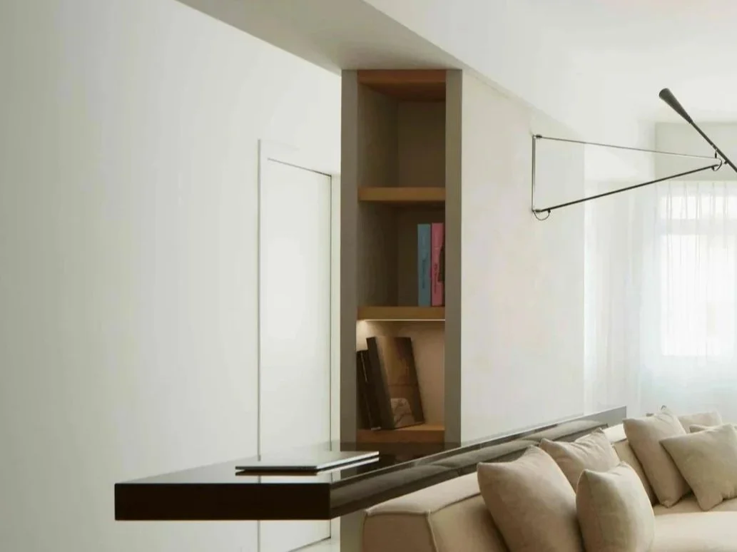 Living room with beige couch, built-in wooden shelves with books, and a black wall-mounted light fixture.