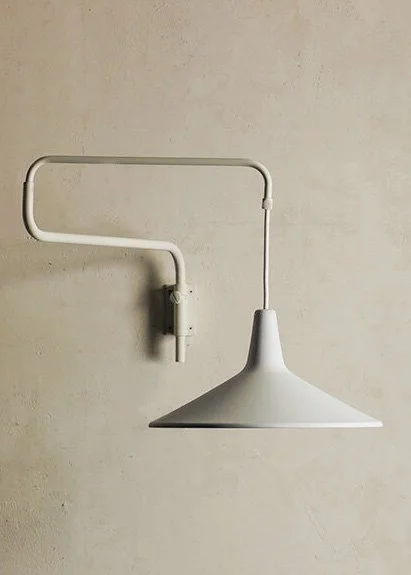 A white wall-mounted lamp with a conical shade and an exposed pipe arm against a beige wall.