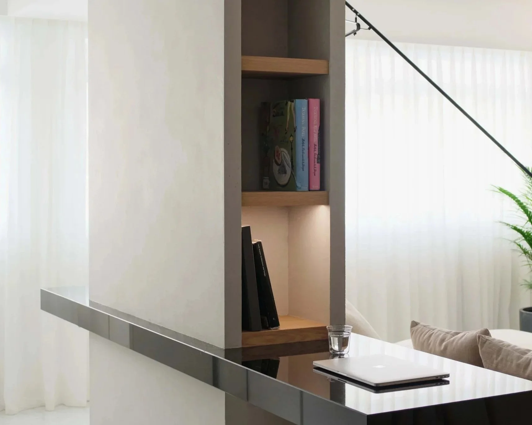 Open black glossy kitchen counter with a laptop, a glass of water, a pillow, and a bookshelf in the background with books and artwork, and sheer white curtains covering large windows.