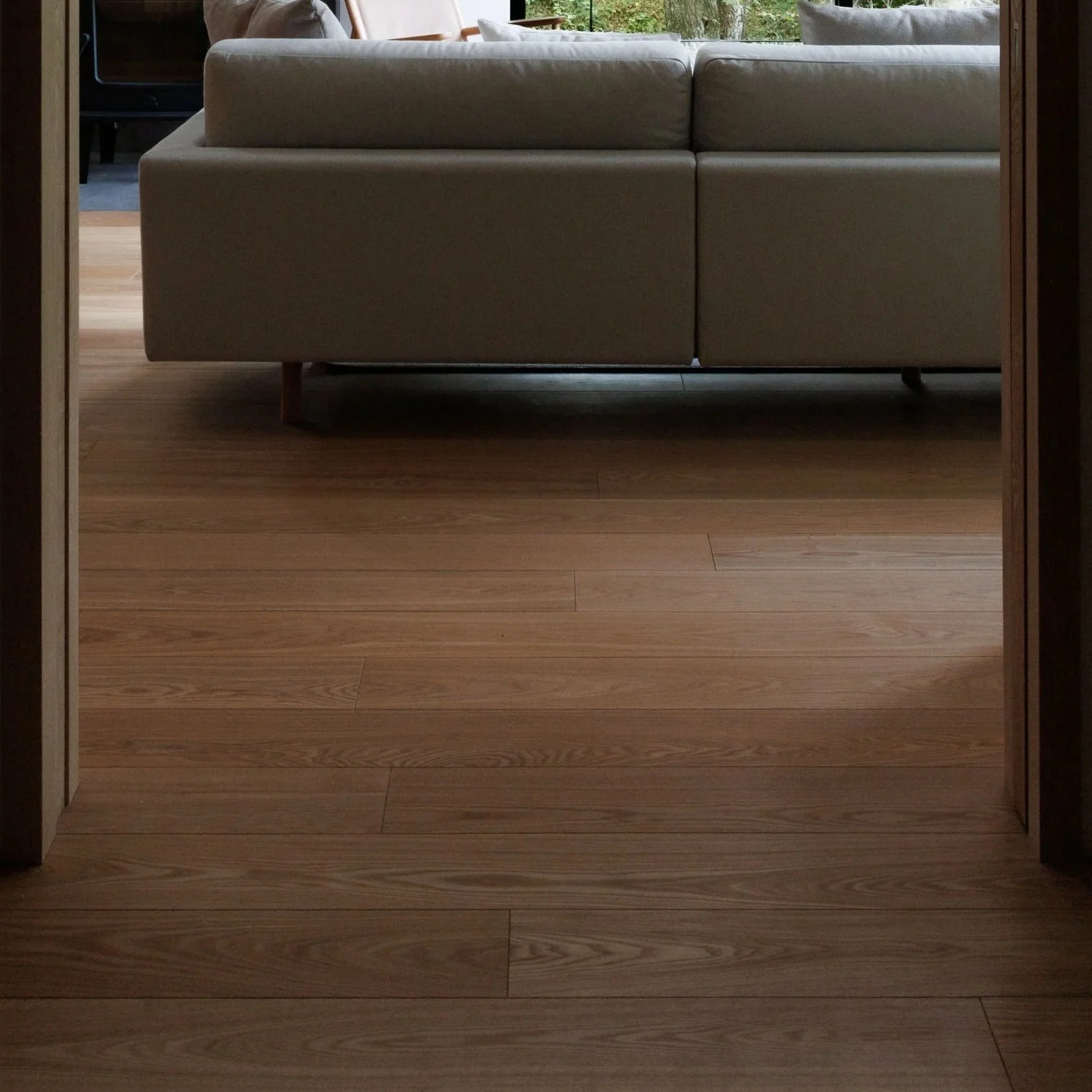 View of a modern living room with a beige sofa on wooden flooring, seen through a doorway with wooden framing. There are cushions on the sofa, and a large window with greenery outside is visible in the background.
