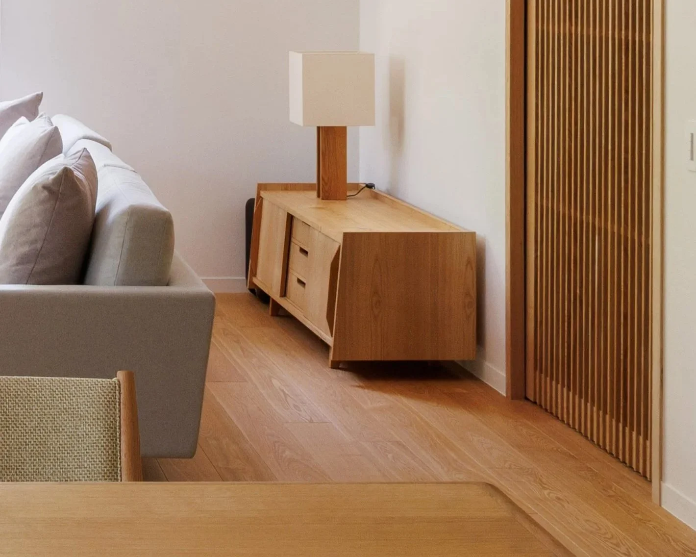 A modern living room corner with a light wood sideboard and a white table lamp, next to a beige sofa with cushions, and a wooden door with vertical slats.