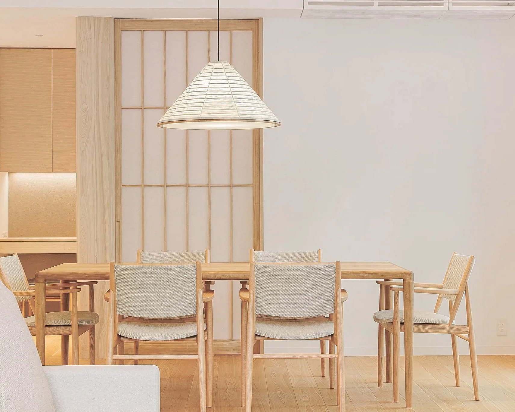 A dining room with a wooden table surrounded by cream-colored upholstered chairs. A large white paper lantern pendant light hangs above the table. The background features a wooden sliding door, a textured wall, and a minimalist interior with light wood flooring.