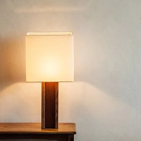 A table lamp with a white shade and a wooden base on a wooden surface against a plain light-colored wall.
