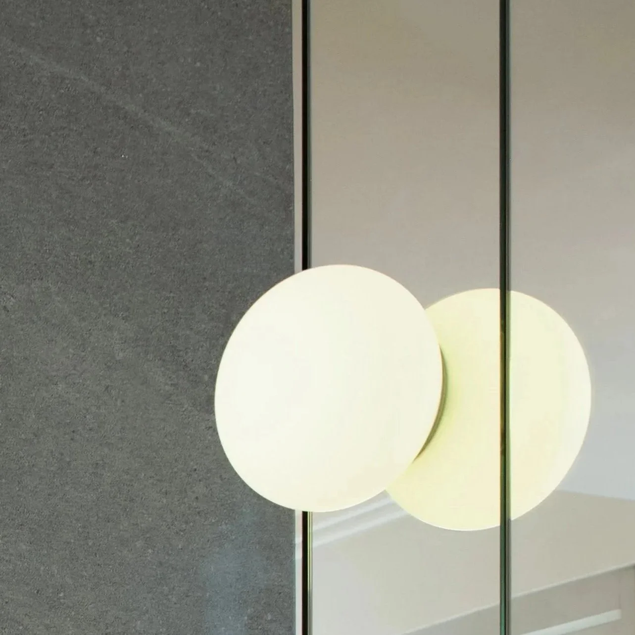 A modern wall-mounted light fixture with two large, round, white globes reflected in a mirror. The background consists of gray and beige wall panels.