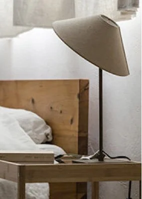 A bedside table with a standing lamp, a book, and a bed with white sheets and a wooden headboard in a minimalist bedroom.