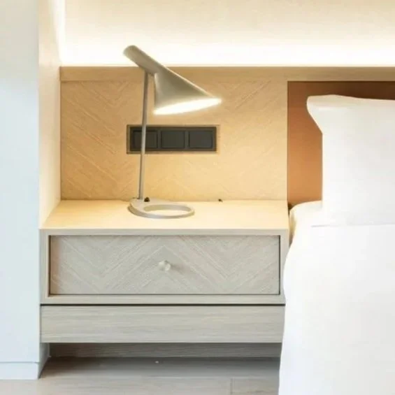 Close-up of a bedside table with a lamp and a bed with white bedding in a modern bedroom.