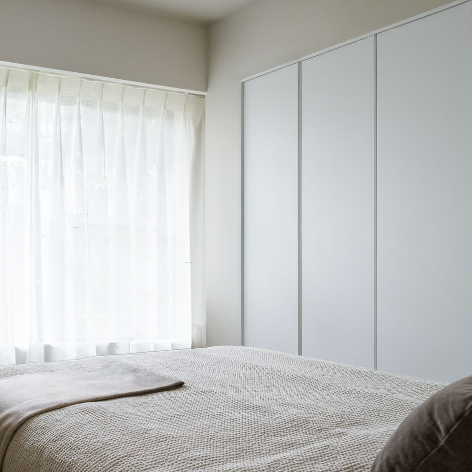 A bedroom with a bed covered in a beige textured blanket, a pillow, a white closet with sliding doors, and a window with white curtains allowing light into the room.