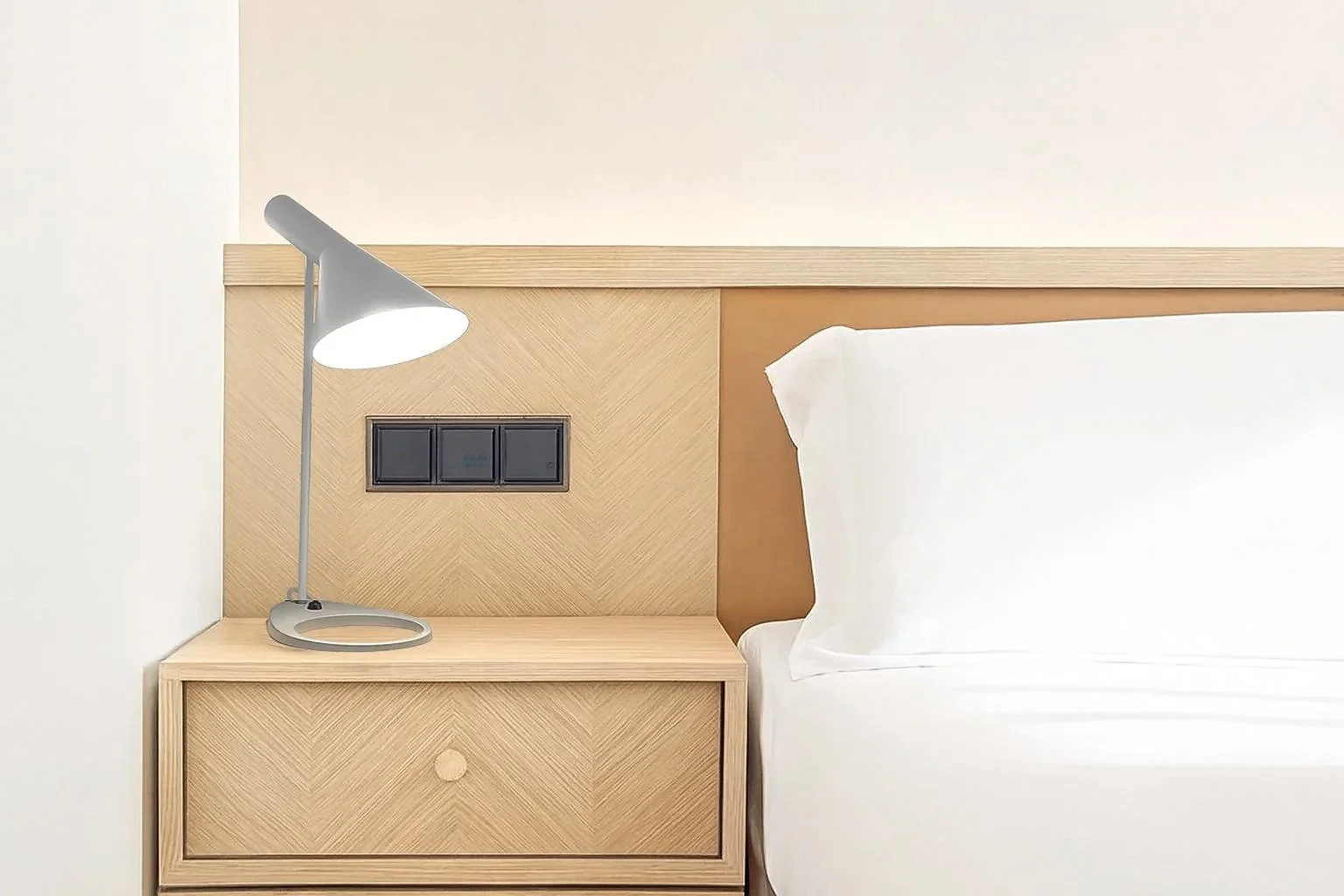 A bedroom nightstand with a light gray desk lamp, adjacent to a bed with white sheets and pillow, against a light-colored wall.