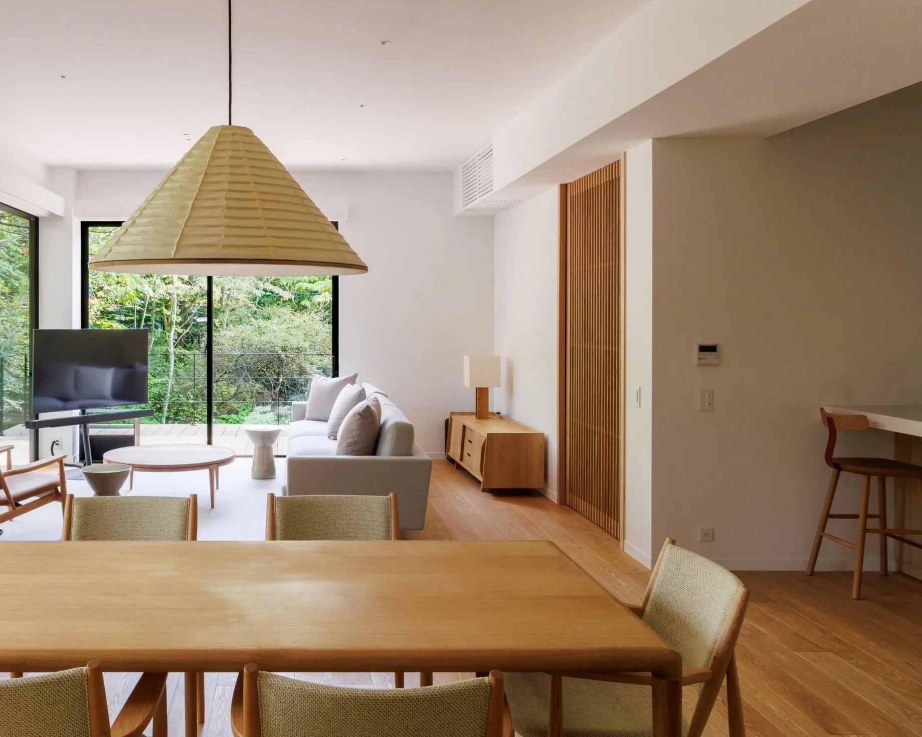 A modern living room with large windows showing greenery outside, a white sofa, a flat-screen TV, wooden furniture including a coffee table and sideboard, a hanging lampshade, and a dining table with chairs.