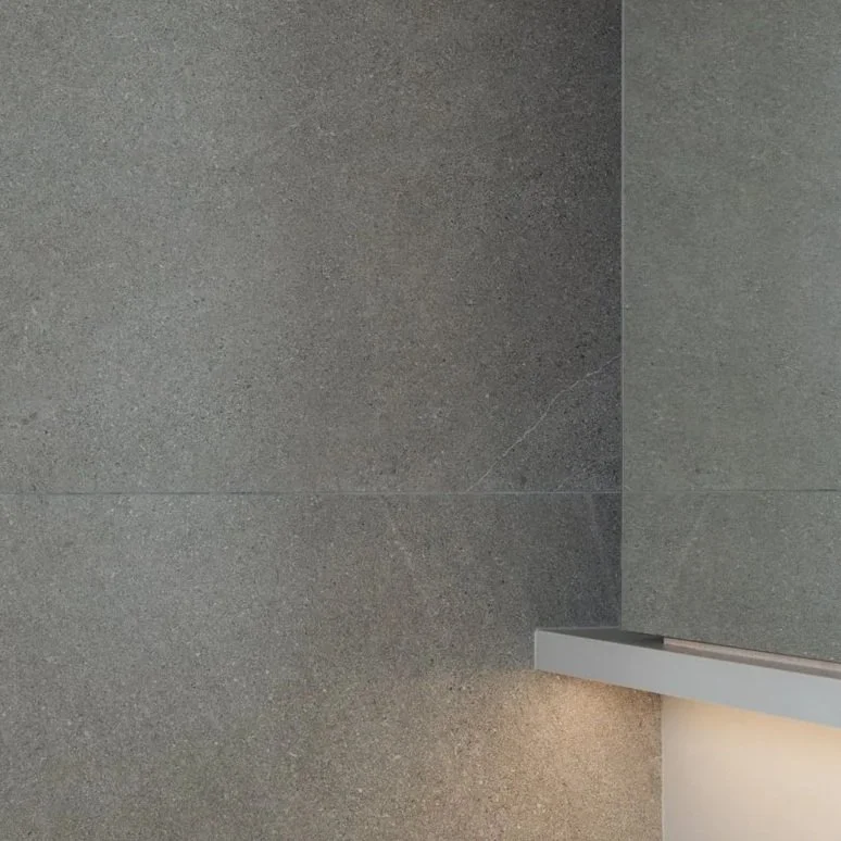 Close-up of gray textured tiled wall and a gray baseboard, with a small warm light at the bottom corner.