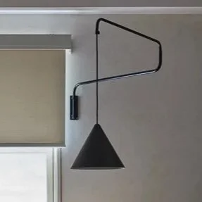 A black wall-mounted desk lamp with an adjustable arm, directed downward, positioned near a window with a beige roller shade.