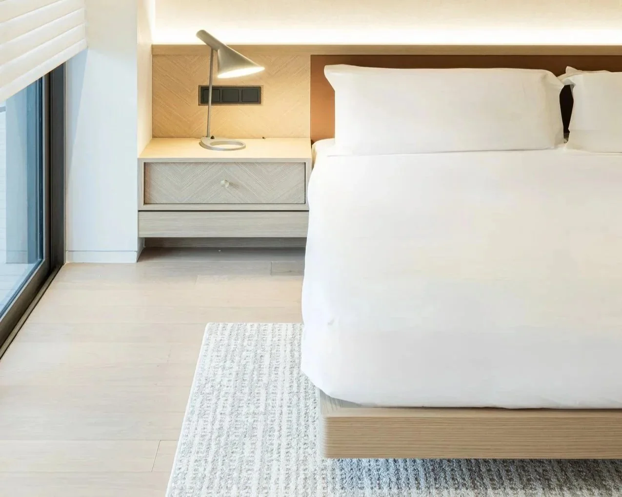 A neatly made bed with white linens and pillows, a wooden nightstand with a modern desk lamp, and a glass sliding door leading to a balcony or patio.