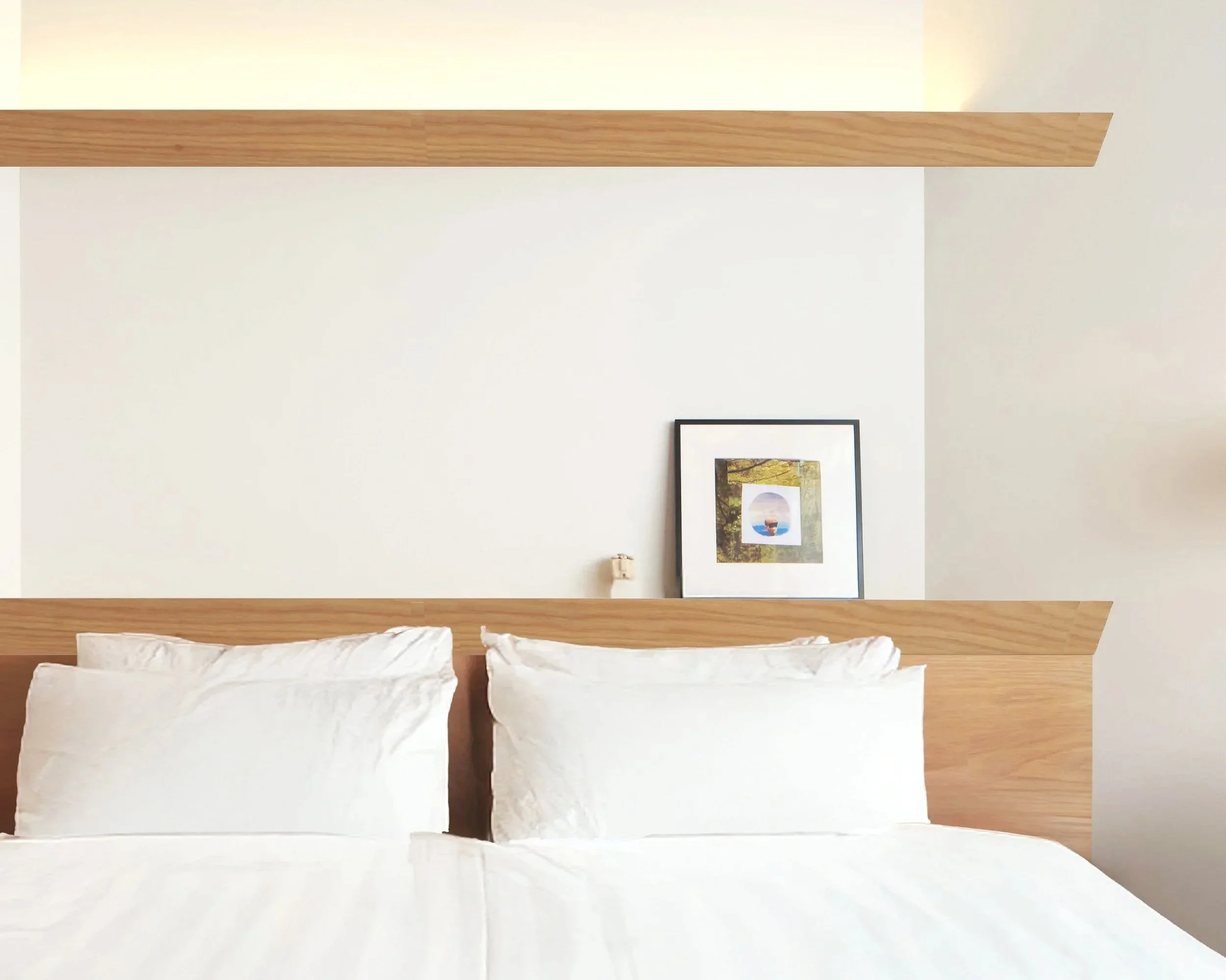 Bedroom with a framed picture hanging on a white wall above a bed with white bedding, and a wooden headboard with a light wood finish.