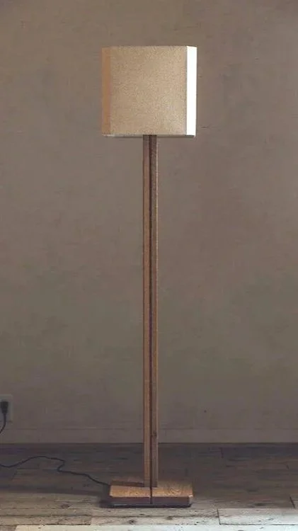 A standing floor lamp with a beige lampshade and a wooden base against a plain wall.