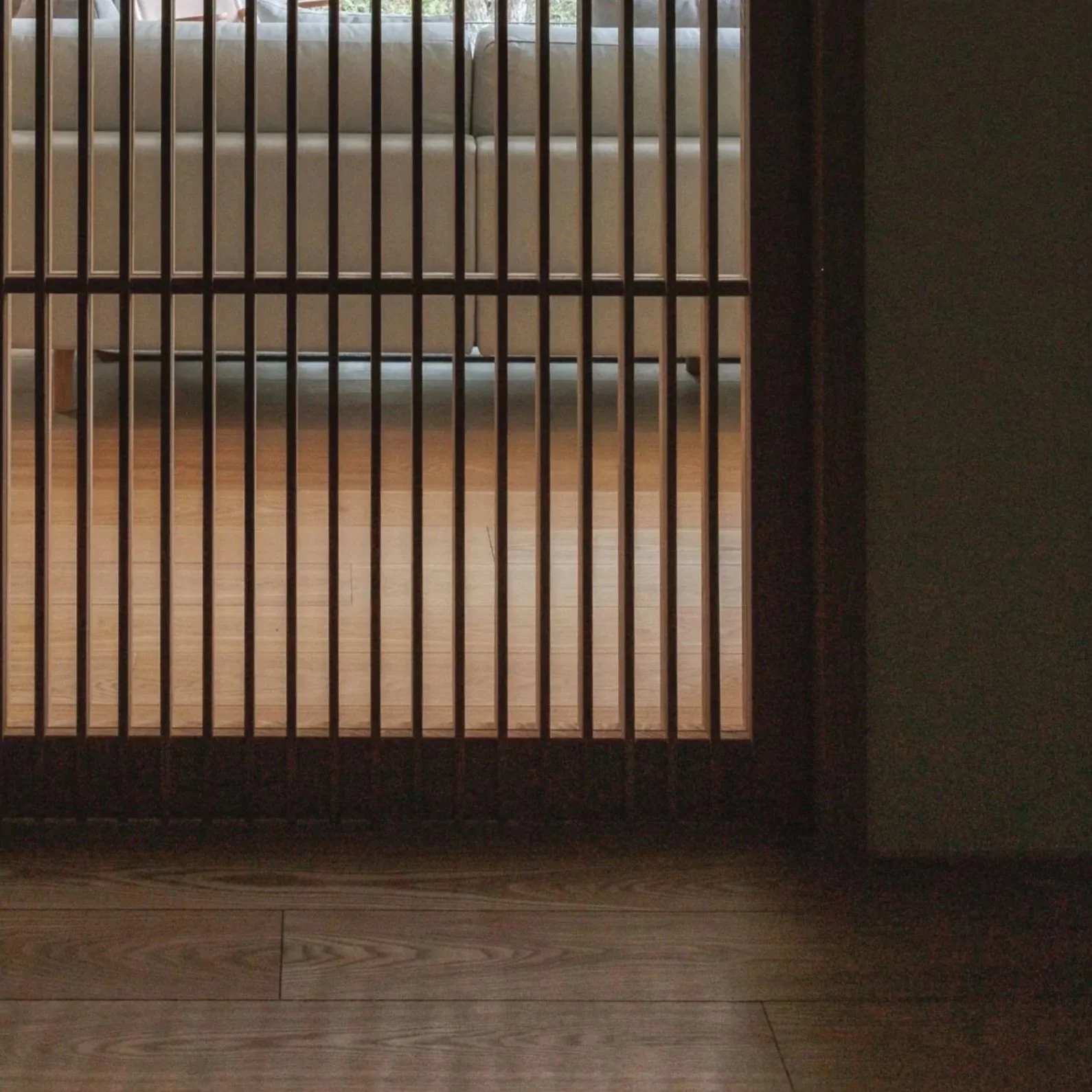 View of a wooden floor behind a black metal gate with vertical bars. Part of a beige sofa with cushions is visible through the bars. The lighting suggests natural light coming from a window or opening behind the sofa.