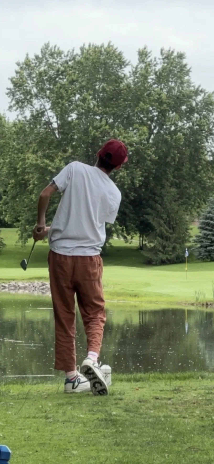 A person playing golf near a water hazard on a golf course, with a flag in the distance and trees in the background.