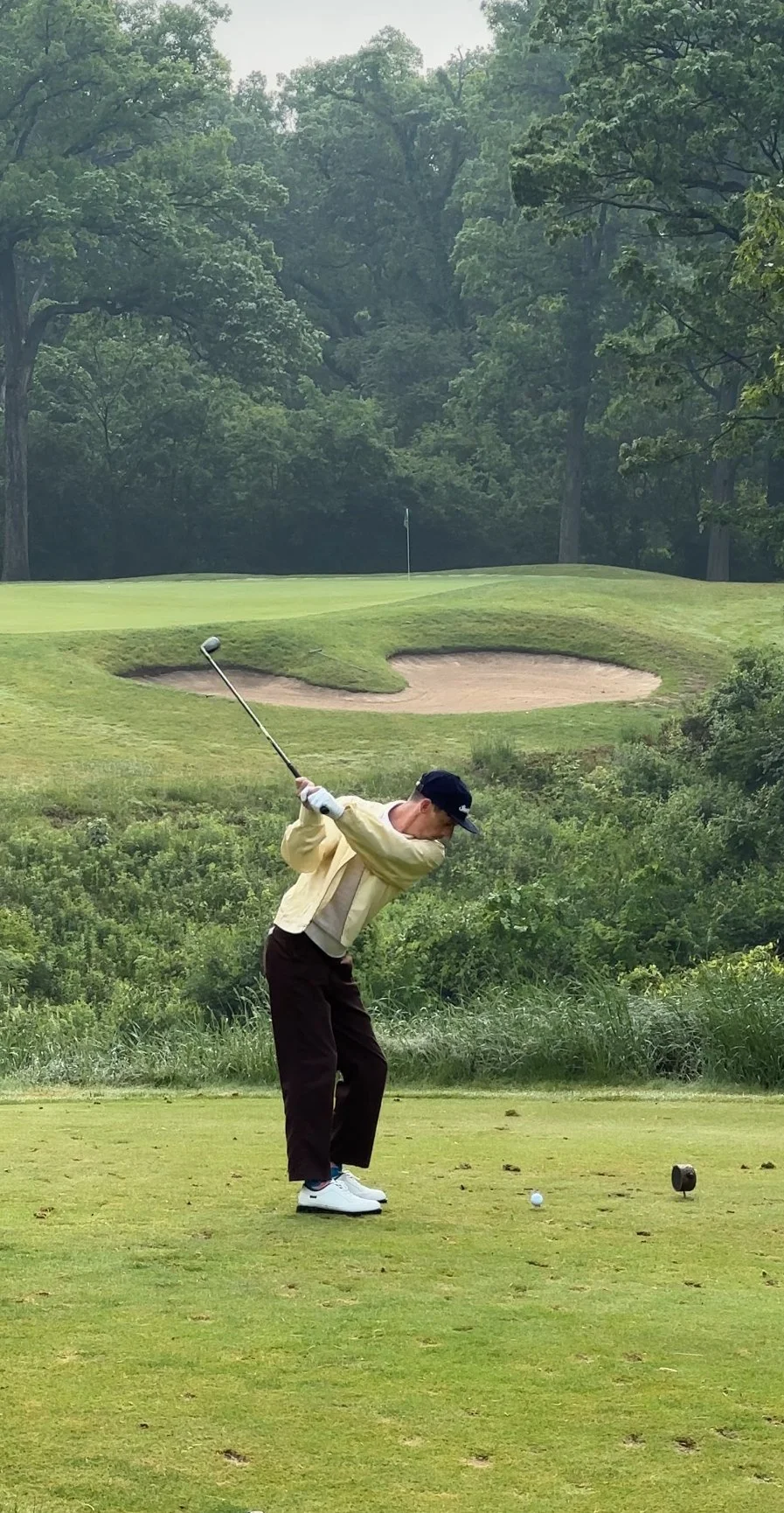 A person wearing a beige jacket, dark pants, white shoes, and a blue cap is swinging a golf club on a golf course, preparing to hit a golf ball. The course has green grass, a sand trap, and a lush backdrop of trees.