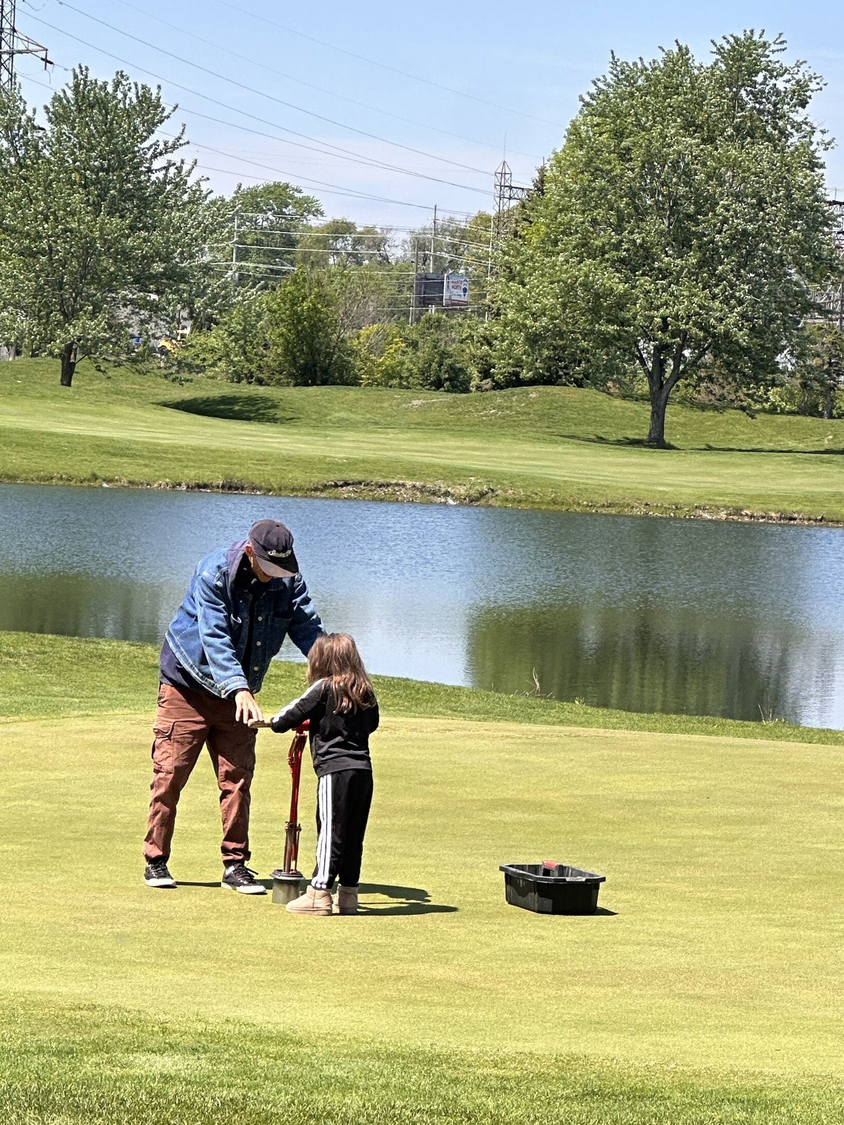 A man teaching a young girl how to use a golf club on a green at a golf course beside a pond, with trees, grass, power lines, and a billboard in the background.