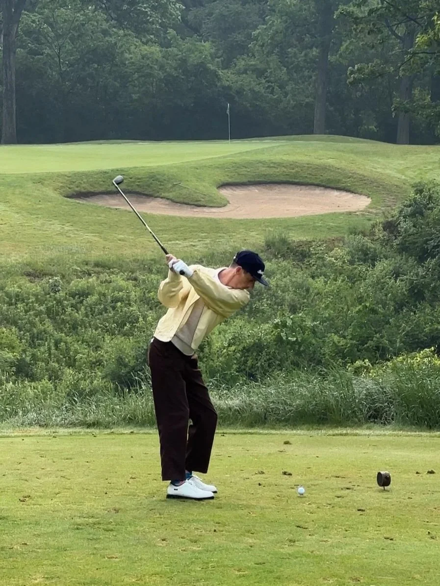 Person playing golf on a course, preparing to hit the ball, with sand bunkers and trees in the background.