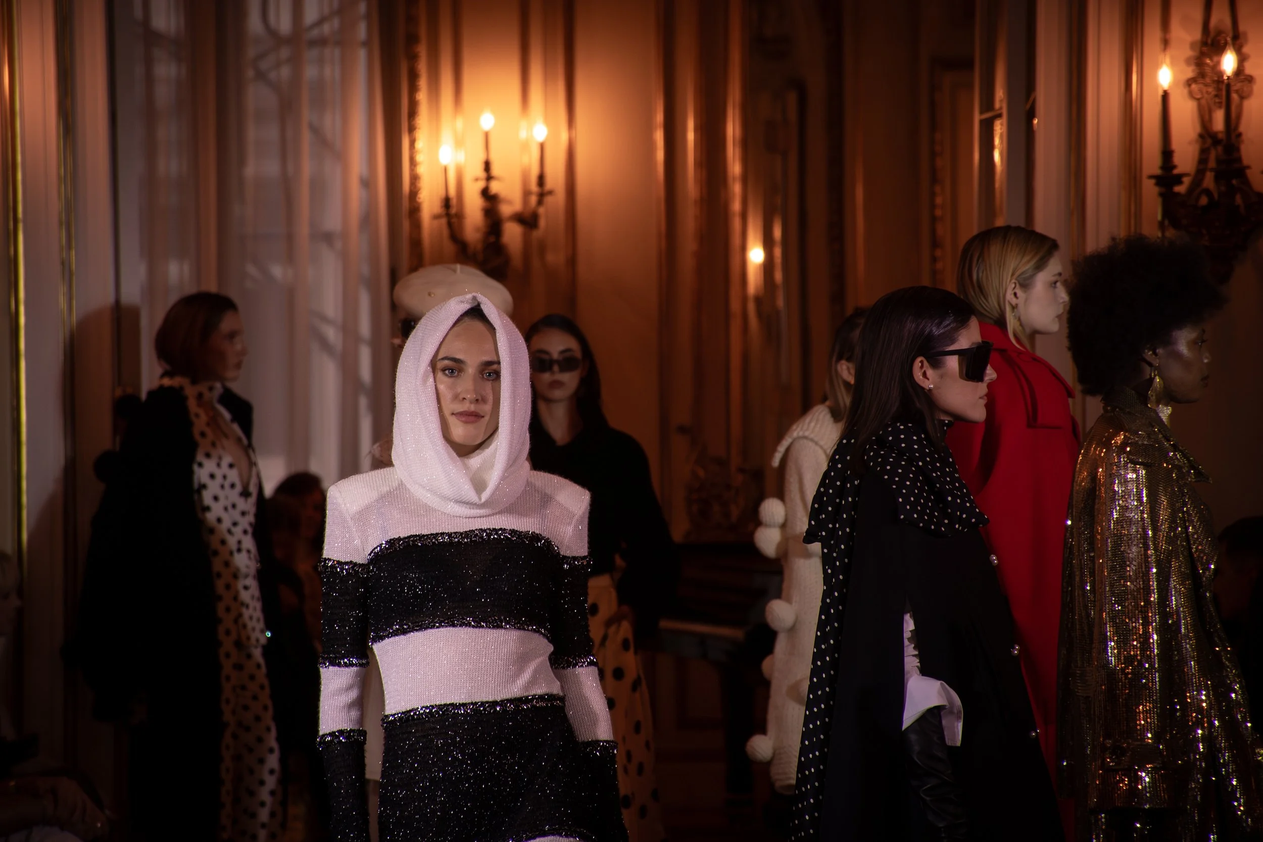 Models standing in a row during a fashion show in a luxurious room with ornate woodwork and chandeliers.