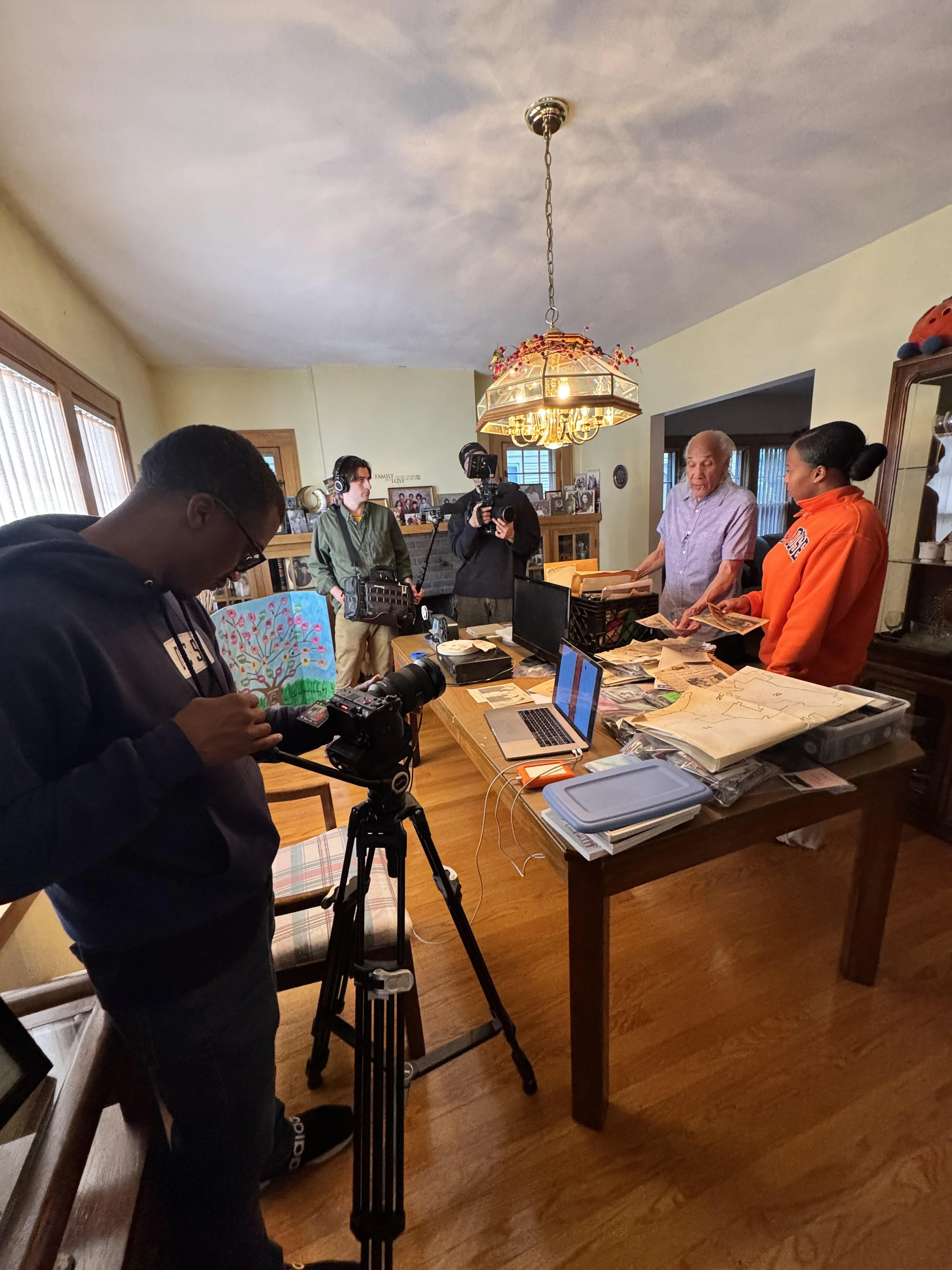 A group of people, including a man with a camera on a tripod, a woman with headphones, an elderly man, and a woman in an orange hoodie, are gathered around a table with papers, photographs, and a laptop inside a warmly lit dining room with light yellow walls, a large chandelier, and family photographs on a fireplace mantel.
