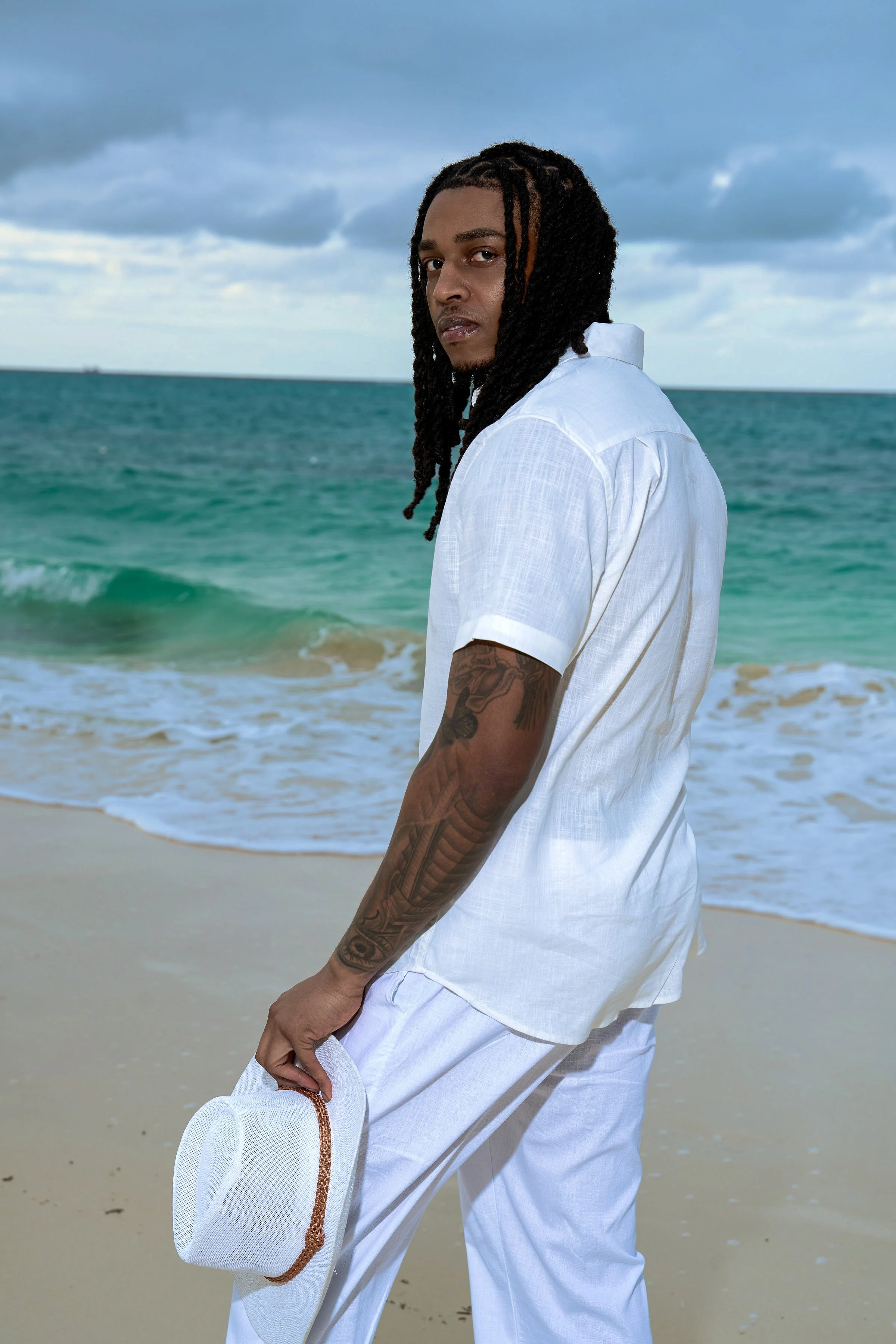 Portrait of a man with long dreadlocks standing on a beach, holding a white hat with a brown band, dressed in white, with the ocean and cloudy sky in the background.