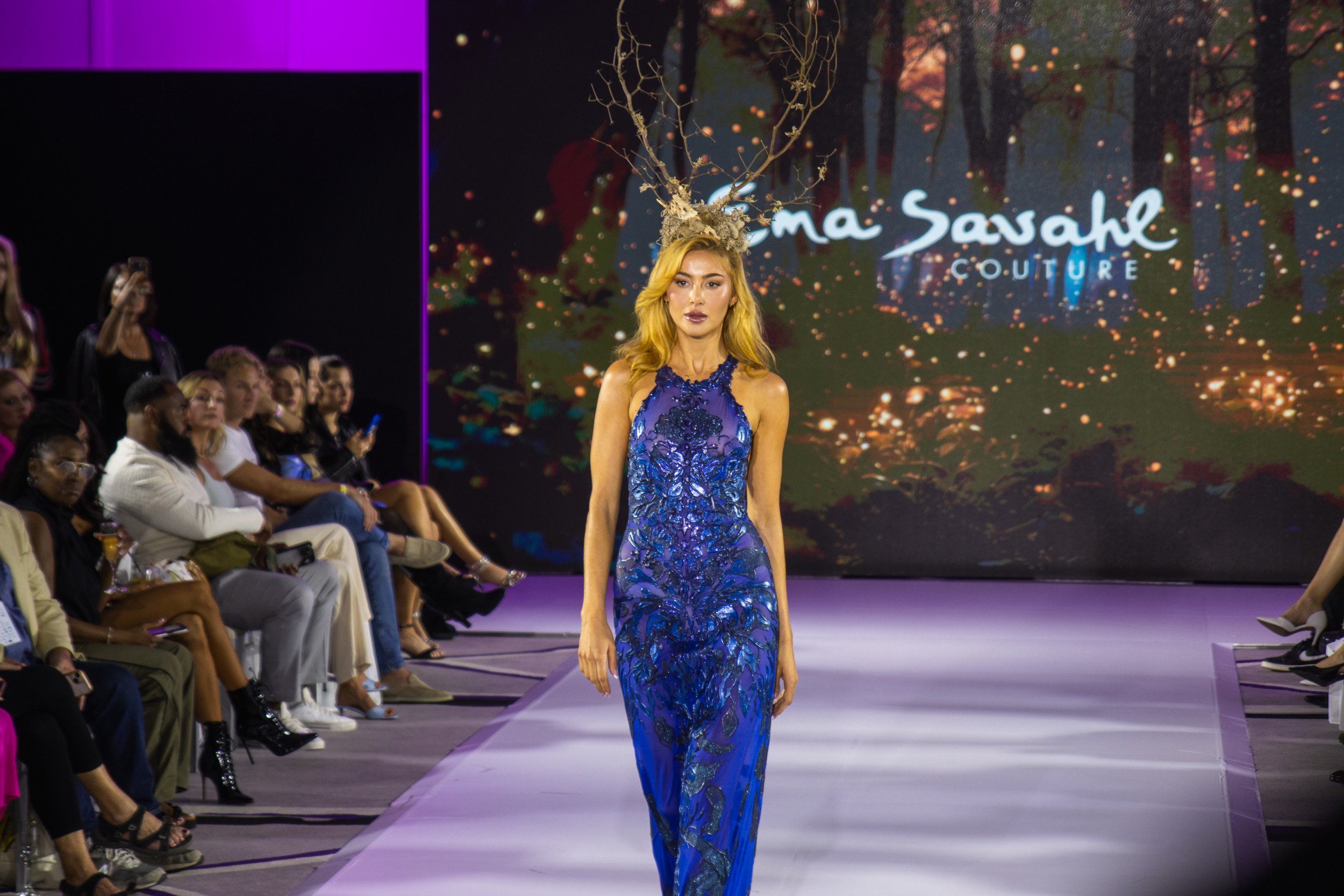 A fashion model with long, wavy blonde hair walks down the runway wearing a blue dress with intricate patterns, and a headpiece resembling a tree with branches and roots, during a fashion show. The background features a large screen displaying the name Emma Savahl and the word Couture, with an abstract forest scene. Audience members are seated on either side of the runway, watching the model.