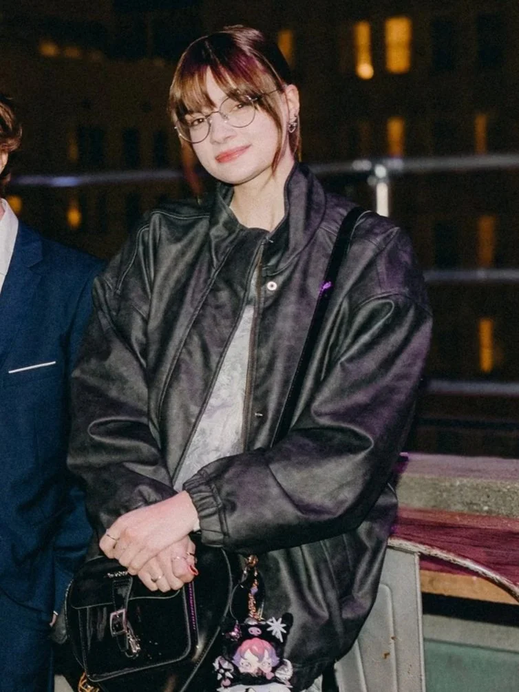 A woman with short brown hair, glasses, and earrings, wearing a black leather jacket, standing outdoors at night. She is holding a small black handbag with a keychain, and there are windows of a building illuminated in the background.