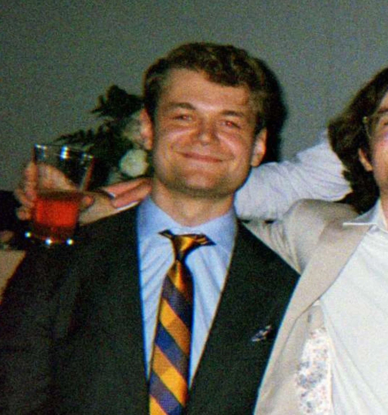 A man with a smile holding a drink in a dark indoor setting, wearing a suit and a colorful striped tie, with another person partially visible next to him wearing a white shirt.