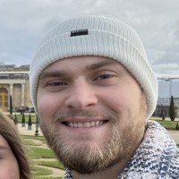 A smiling man wearing a white knit beanie outdoors with a park and water in the background.