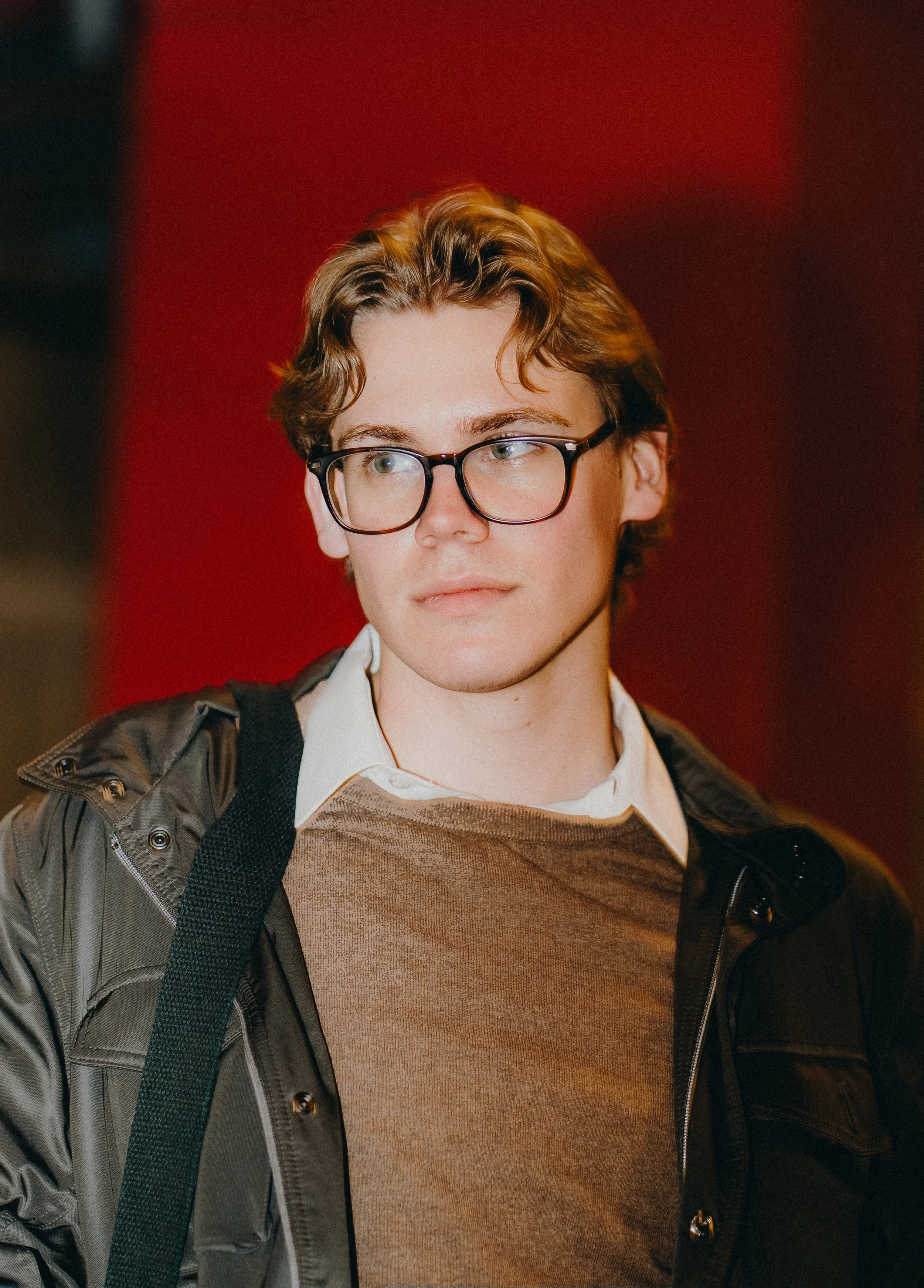 Young person with light skin, curly light brown hair, and glasses, wearing a white shirt and brown sweater, carrying a black backpack, standing against a red background.