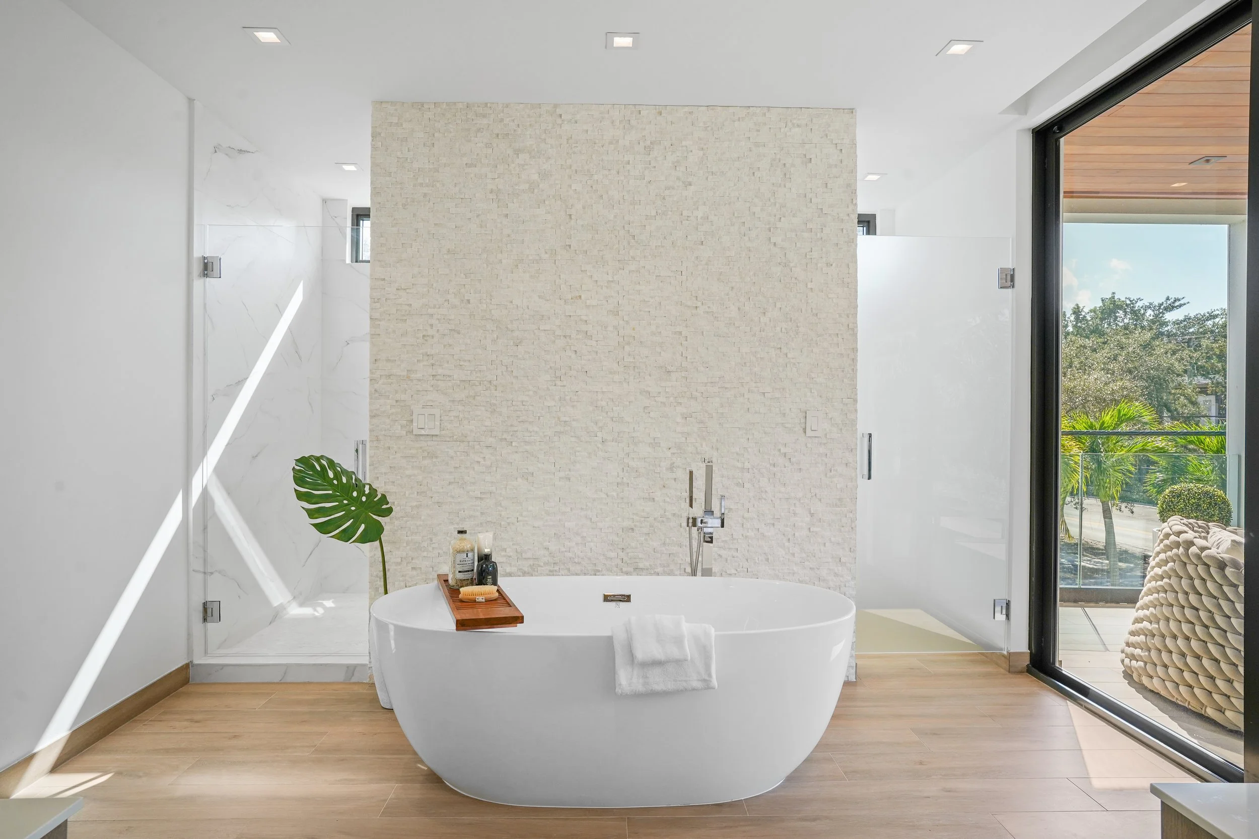Modern bathroom with a white freestanding bathtub, a wooden tray with bath accessories, a potted plant, a textured beige wall, and large windows showing trees outside.