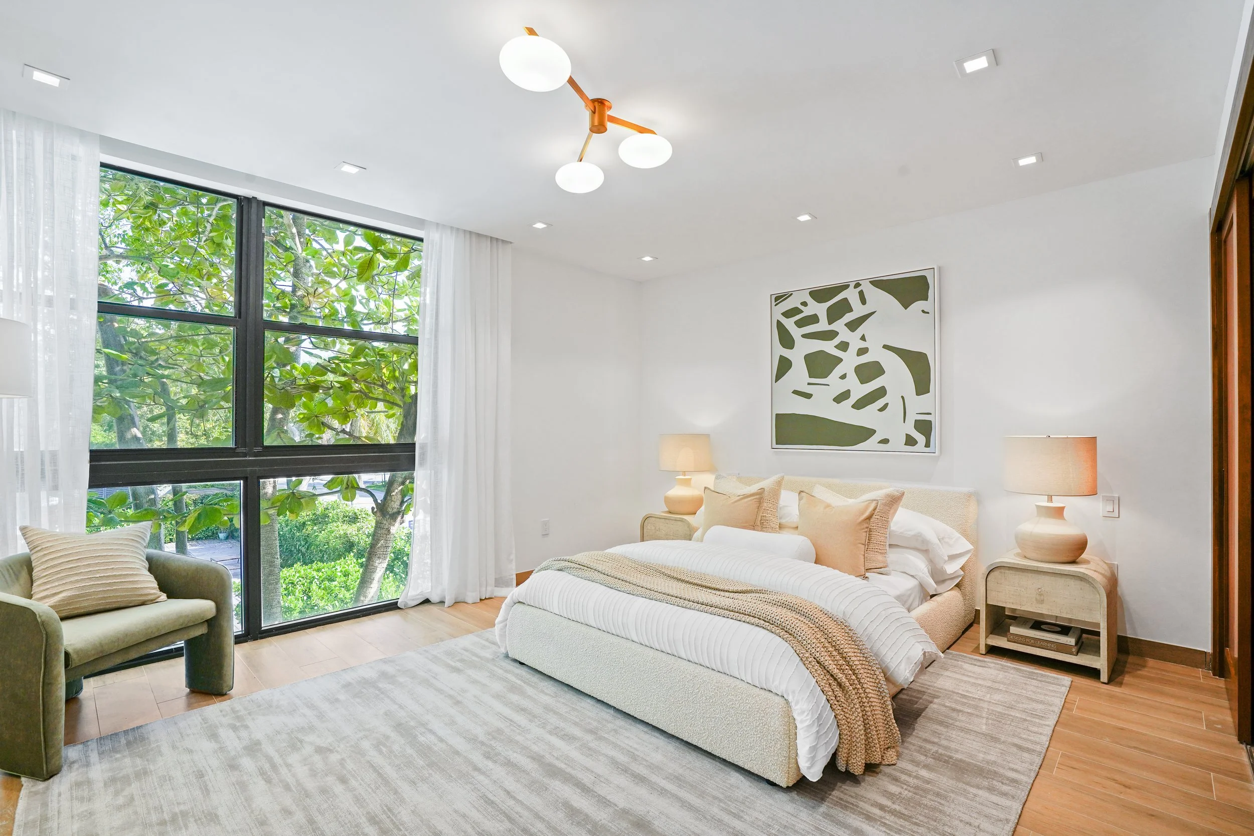 Bright modern bedroom with large window showing green foliage outside, white walls, beige bed with neutral bedding, two matching beige nightstands with lamps, abstract artwork on the wall, and a small sofa with striped pillows.