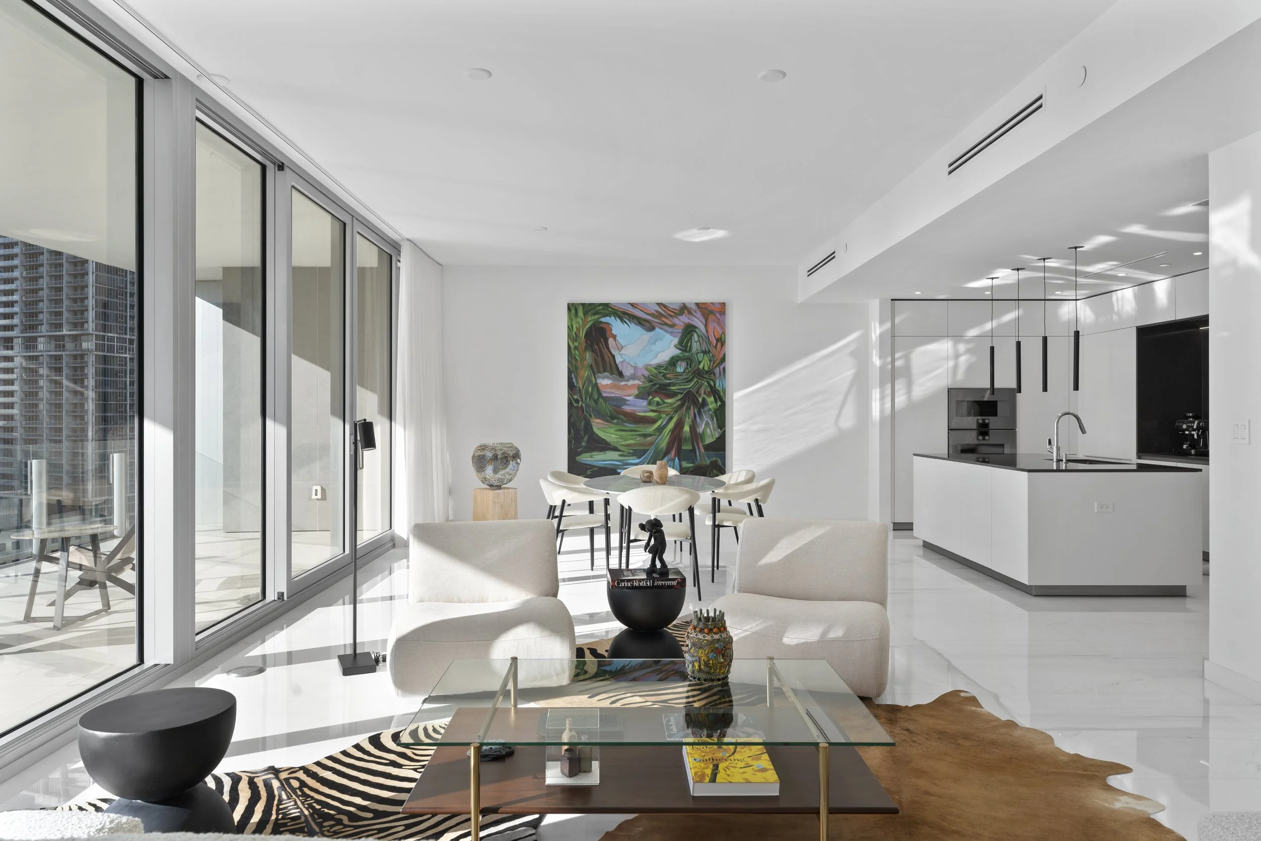 Modern open-concept interior with white walls and large glass sliding doors leading to balcony, featuring a living and dining area with abstract art, decorative objects, black and white furniture, and natural sunlight streaming through.
