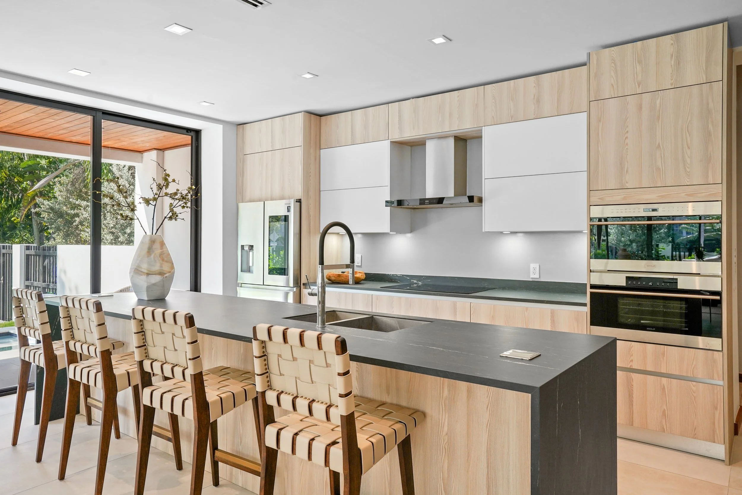 Modern kitchen with beige and white cabinetry, black countertop island with a sink, woven-wrapped chairs along the island, oven, refrigerator, and sliding glass door leading outside to greenery.
