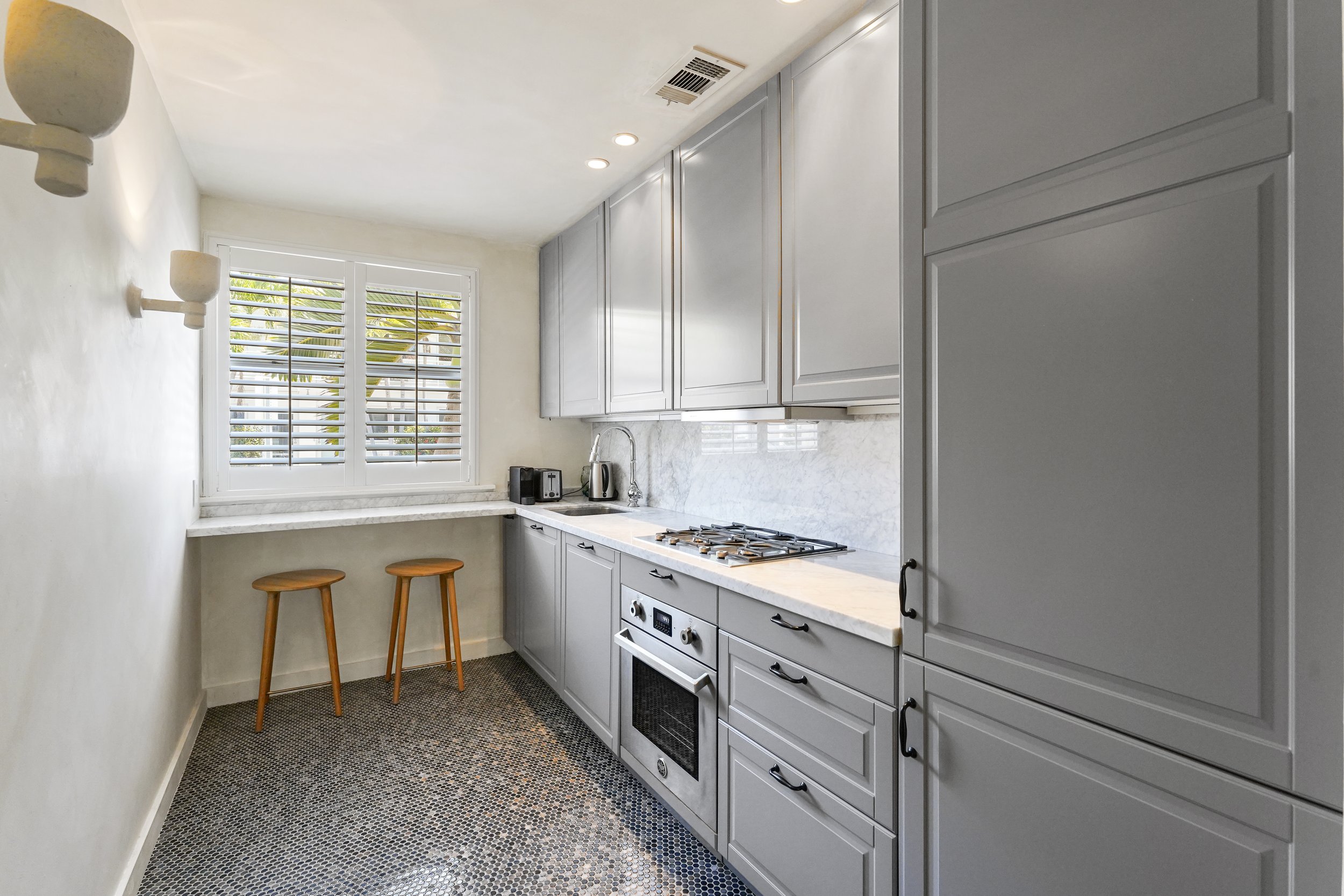 A small kitchen with light gray cabinets, a stove, and a marble countertop, with a window with white shutters and two wooden bar stools.