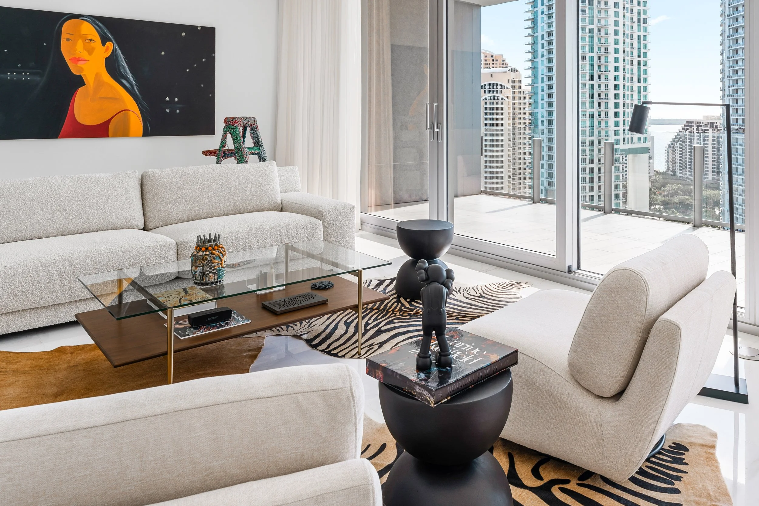 Living room with white sofas, glass coffee table, abstract black and gray side tables, artwork of a woman with long dark hair, city view through large windows, balcony, modern high-rise buildings, floor lamp, zebra pattern rug.