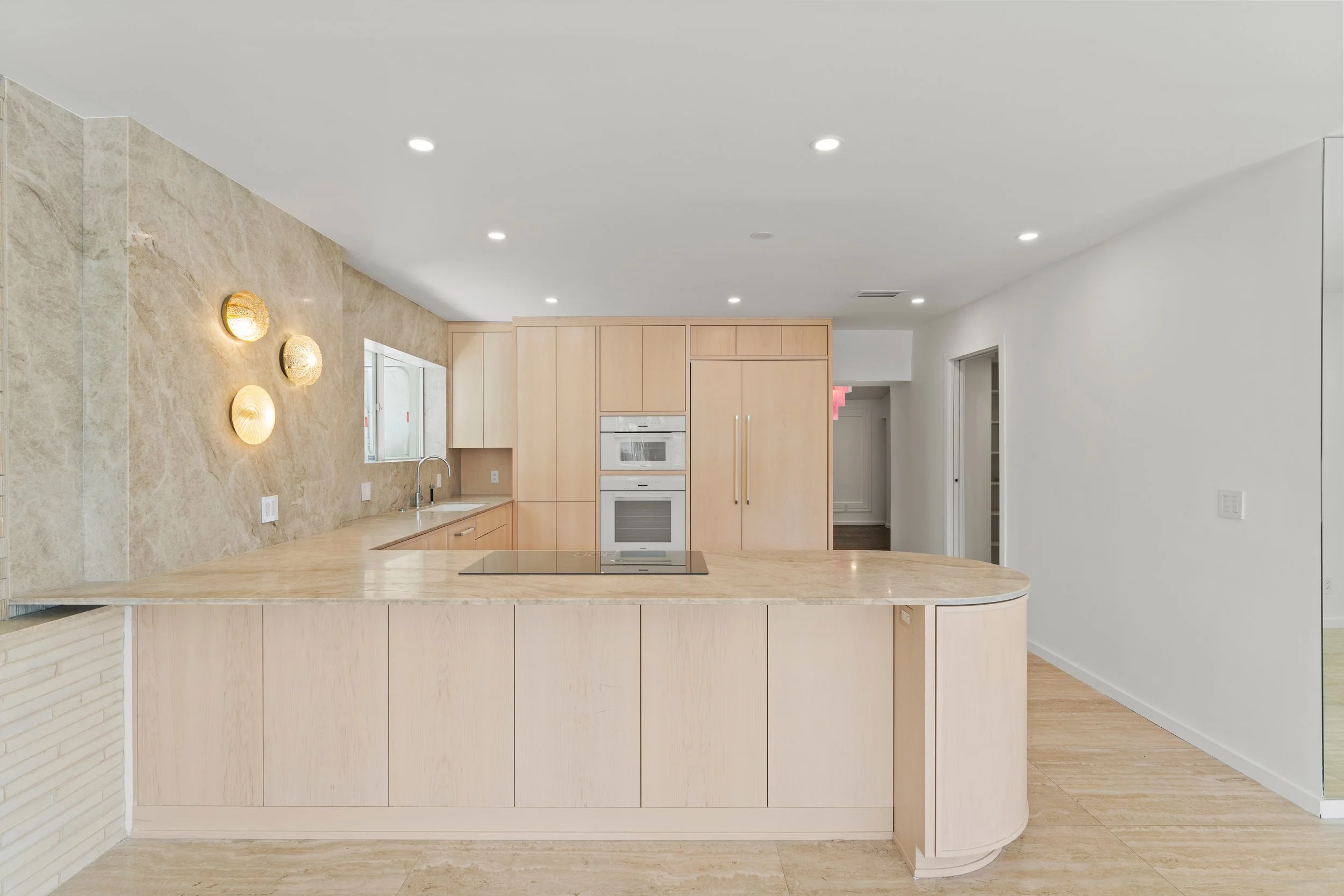 Modern kitchen with light wood cabinets, marble countertops, a built-in oven, and decorative wall lights.