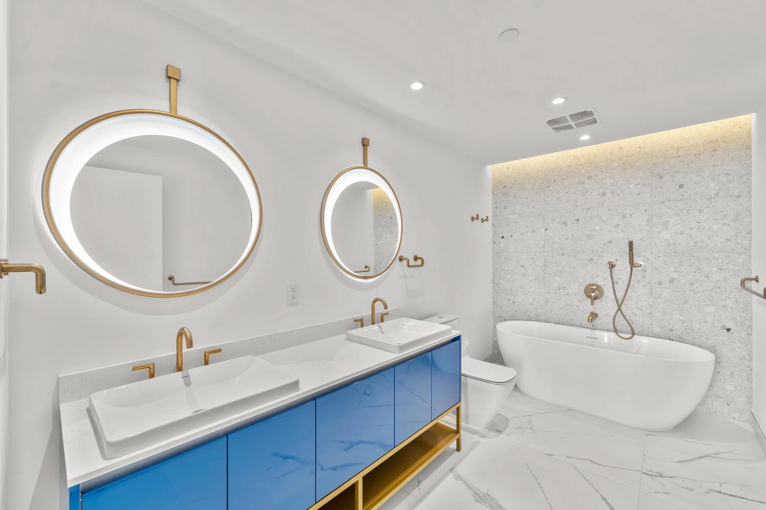 Modern bathroom featuring two round illuminated mirrors, a white dual sink vanity with blue cabinets and gold accents, a freestanding bathtub, and a shower area with stone wall tiles and gold fixtures.