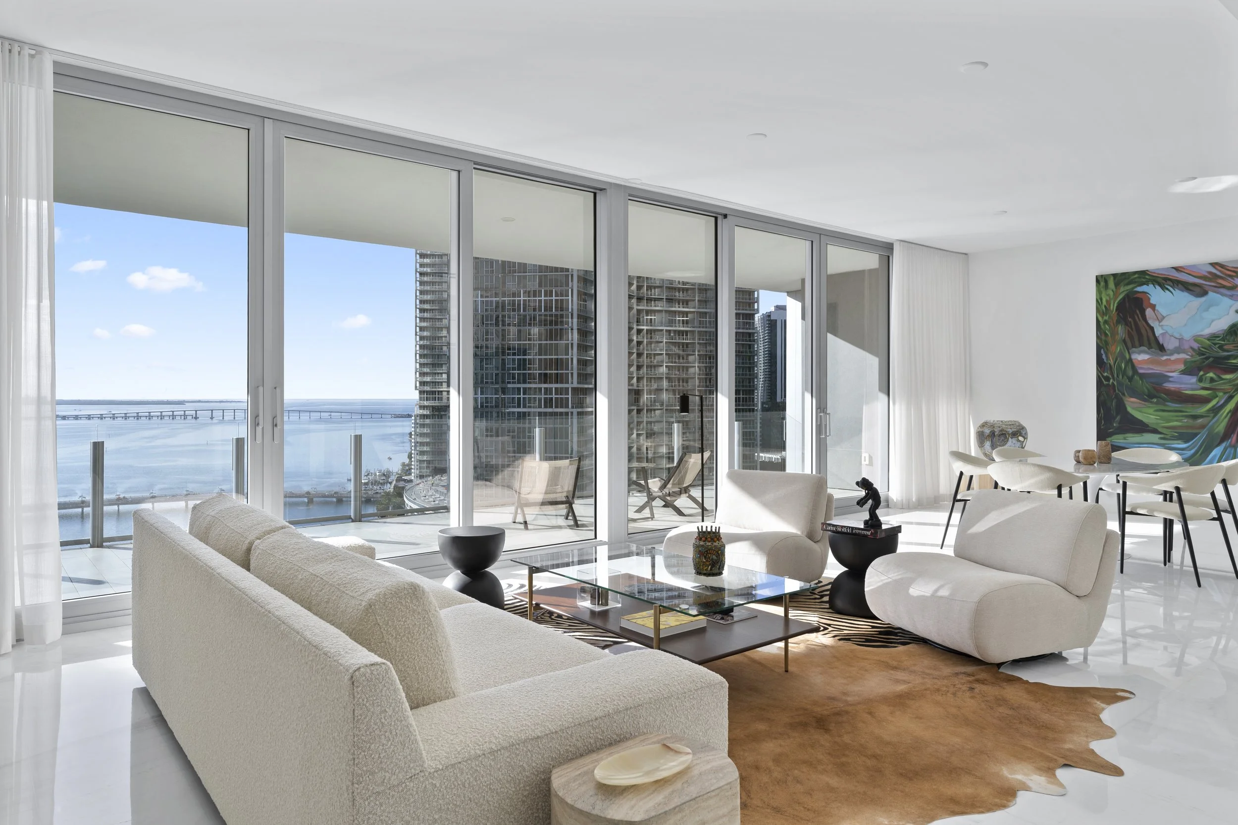 Modern living room with large floor-to-ceiling windows overlooking a city skyline and water, white sofas, a glass coffee table, and colorful artwork on a white wall.