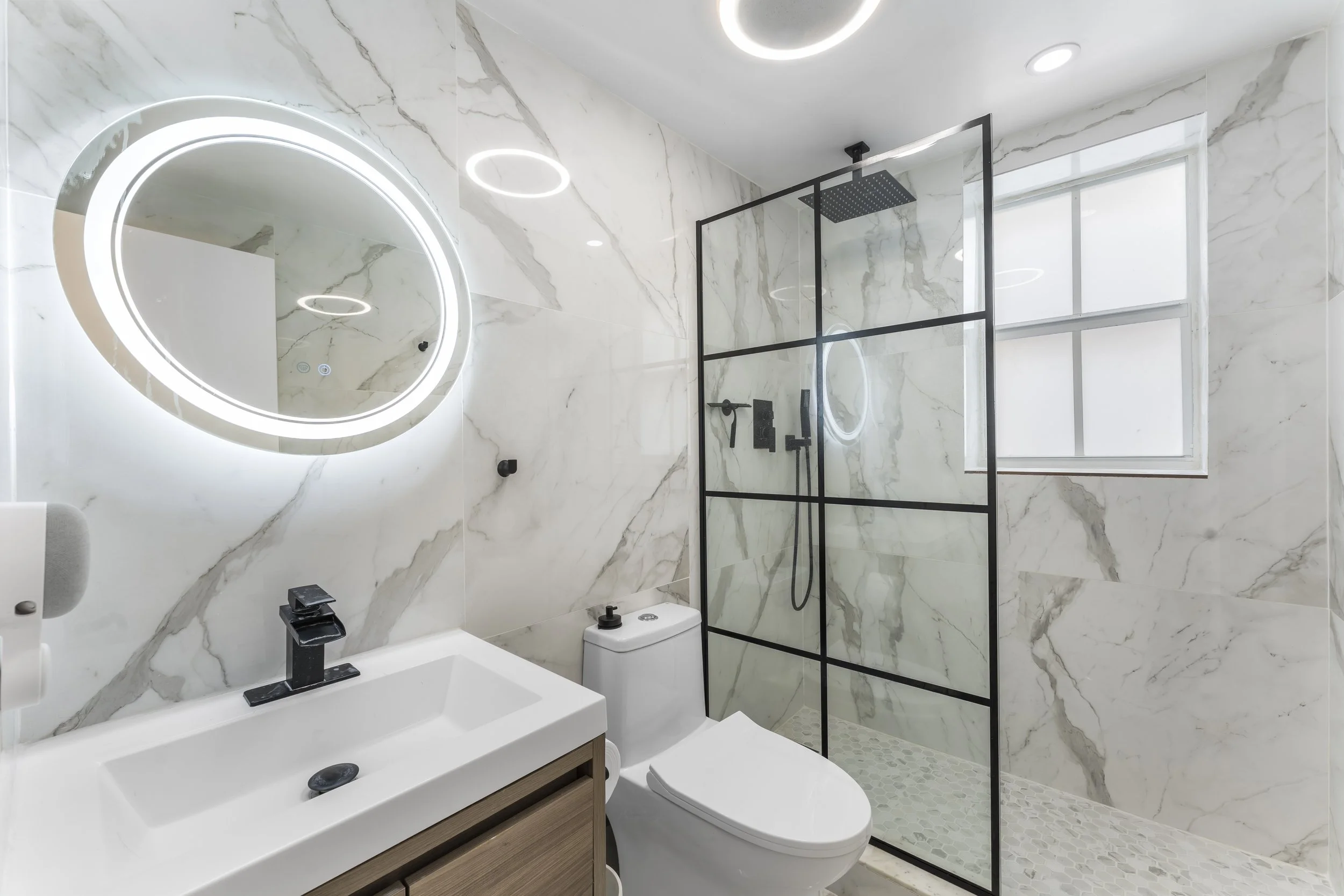 Modern bathroom with marble walls and a large mirror with LED lighting. Features include a black shower frame with rainfall showerhead, a white toilet, and a vanity with a white sink, black faucet, and wooden cabinet.