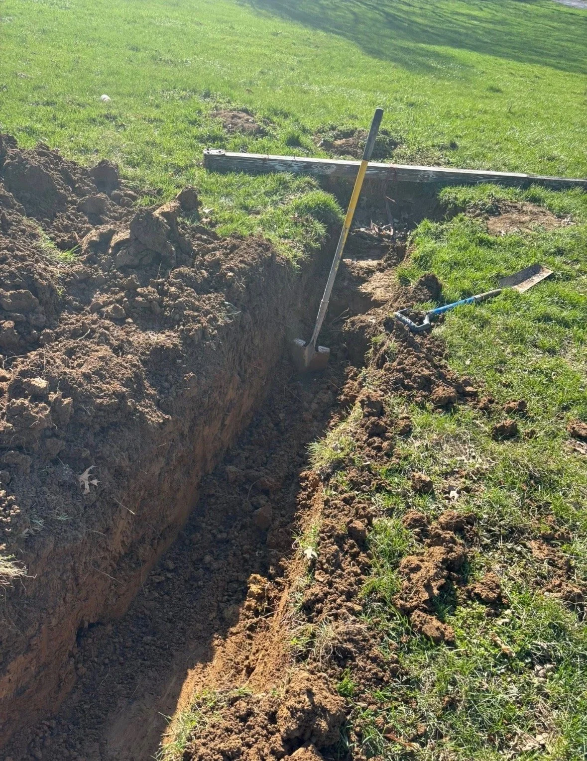 A deep trench in the ground with tools, a shovel, and a measuring stick nearby, with a wooden board across the top of the trench.