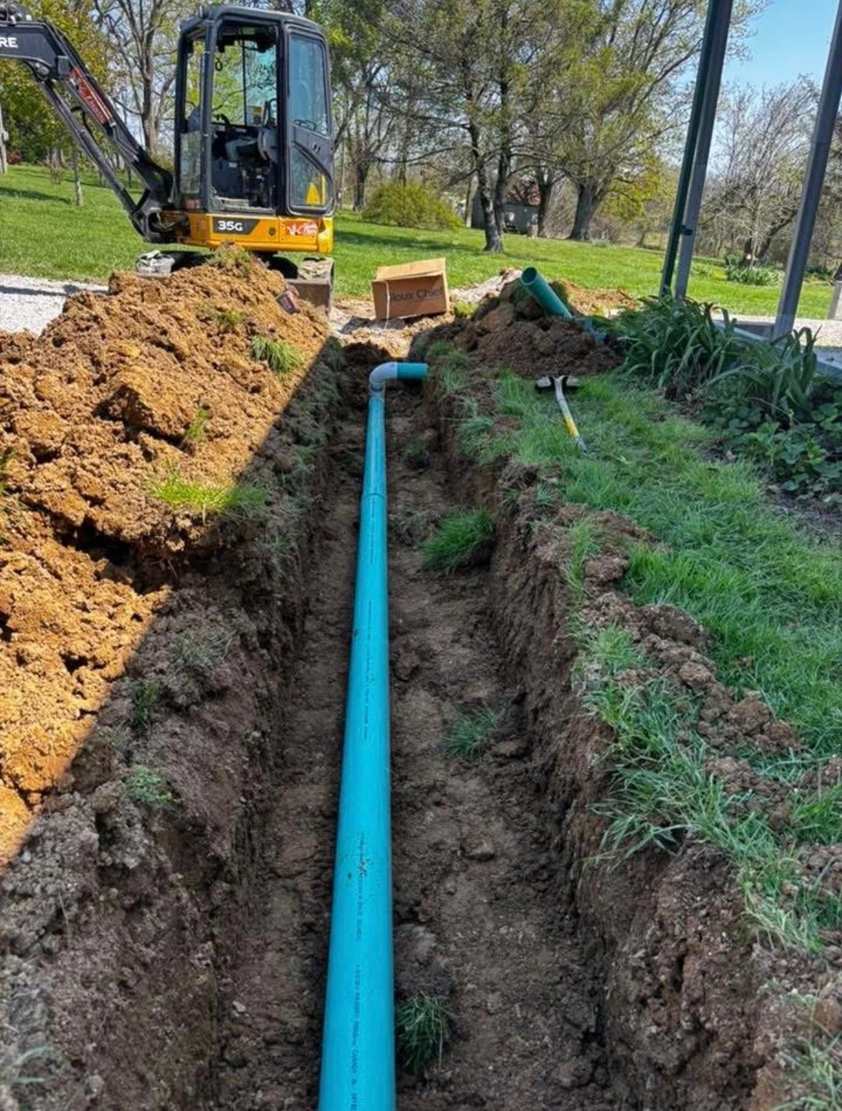 An outdoor scene shows a blue underground pipe installed in a freshly dug trench, with a small excavator and construction tools nearby, in a grassy yard with trees.