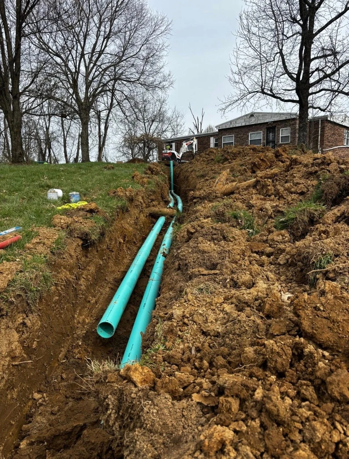 Two blue pipes installed in a trench in front of a brick house with leafless trees on a cloudy day.